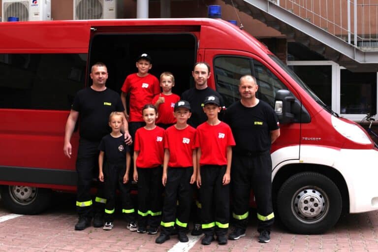 Grupa dzieci i dorosłych reprezentująca Straż Pożarną pozuje do pamiątkowej fotografii. Dzieci ubrane są w czerwone koszulki i spodnie strażackie. Pozują na tle czerwonego busa strażackiego