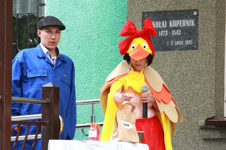 Od lewej mężczyzna w niebieskim kombinezonie i czapce robotniczej, obok kobieta w przebraniu kaczki, w stroju żółto pomarańczowym sięga po brązową torbę z napisem: mak