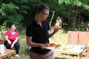 W centrum kadru młodzieniec w czarnej koszulce, beżowych spodniach i okularach. W prawej ręce trzyma talerz z pizzą, w lewej kawałek pizzy. W tle zieleń