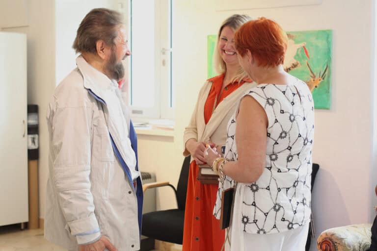 Na fotografii trzy osoby pogrążone w rozmowie. Kobieta, która stoi przodem do obiektywu, śmieje się