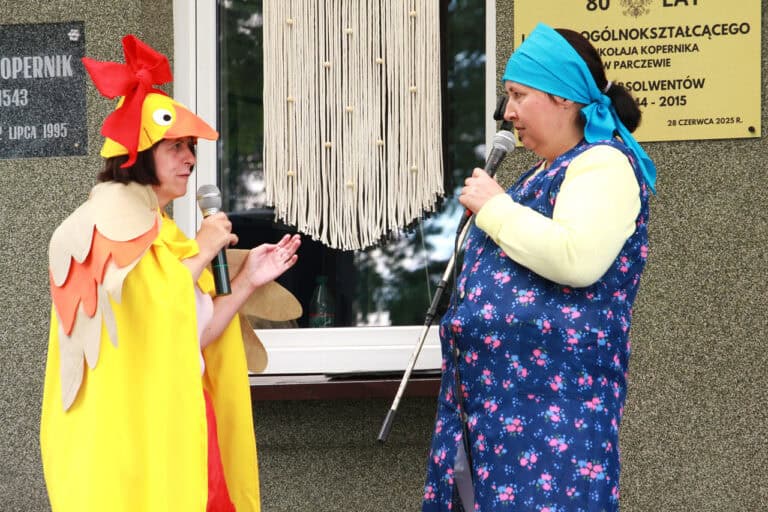 Od lewej kobieta w przebraniu kaczki, z dużą czerwoną kokardą na głowie zwrócona w stronę kobiety ubranej w kwiecisty fartuch, z niebieską chustką na głowie