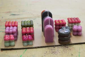 Prace artystyczne uczestników warsztatów: Świece na tekturowej podkładce. Są w barwach różu, czerwieni i zieleni. Owalne i prostokątne