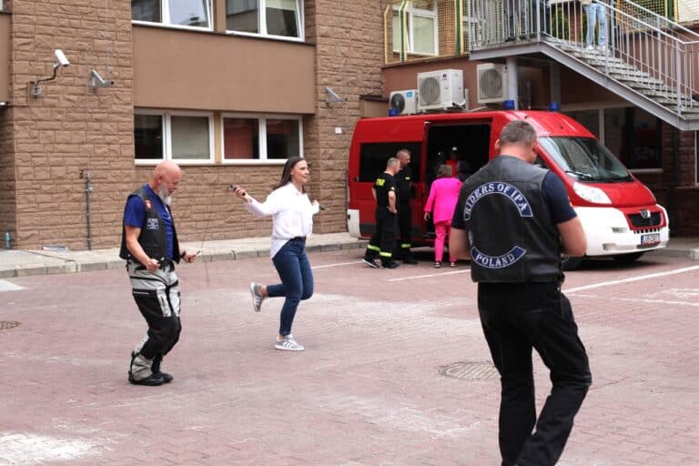 W centralnej części kadru dwie osoby skaczące na skakankach. Jedną z nich (kobieta) jest dyrektor placówki. W tle wóz strażacki i ściana budynku szpitala