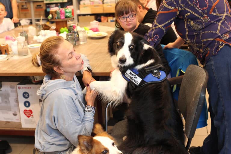 W centralnej części fotografii prowadząca zajęcia i siedzący na krześle pies wykonujący komendę „podaj łapę”. Pies ogląda się w stronę aparatu. Ma na sobie szelki z napisem: Megi, pies terapeuta