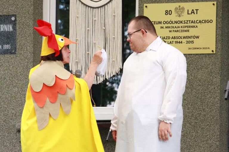 Od lewej kobieta w stroju kaczki, w barwach żółtych i pomarańczowych pokazuje mężczyźnie, który stoi przed nią mały biały woreczek. Mężczyzna ma na sobie biały fartuch i okulary, uśmiecha się