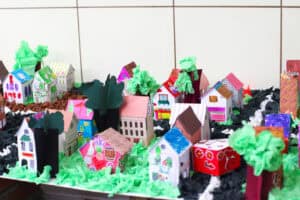 Makieta miasta, wykonana z kartonów o różnych kolorach: różowe, czarne, pomarańczowe, niebieskie, zielone i żółte. Na makiecie znajdują się domy i sklepy i drzewa