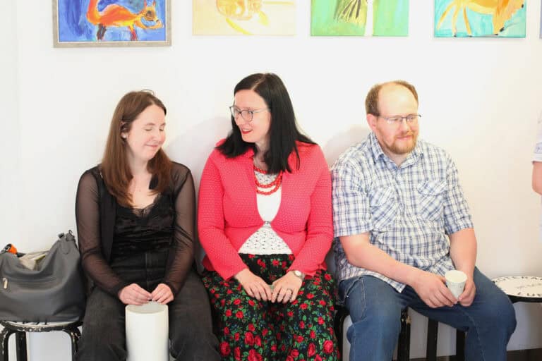 Trzy osoby siedzą na taboretach: dwie kobiety i mężczyzna. Kobiety zwracają się ku sobie, rozmawiając. Mężczyzna uśmiecha się. Nad nimi widać fragmenty obrazów