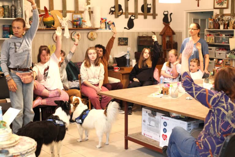 Kadr wypełniony młodzieżą i opiekunami zajęć zgromadzonymi wokół stołu i na kanapach podczas dogoterapii. Prawie wszyscy unoszą w górę lewą rękę. W lewym dolnym rogu kadru dwa psy