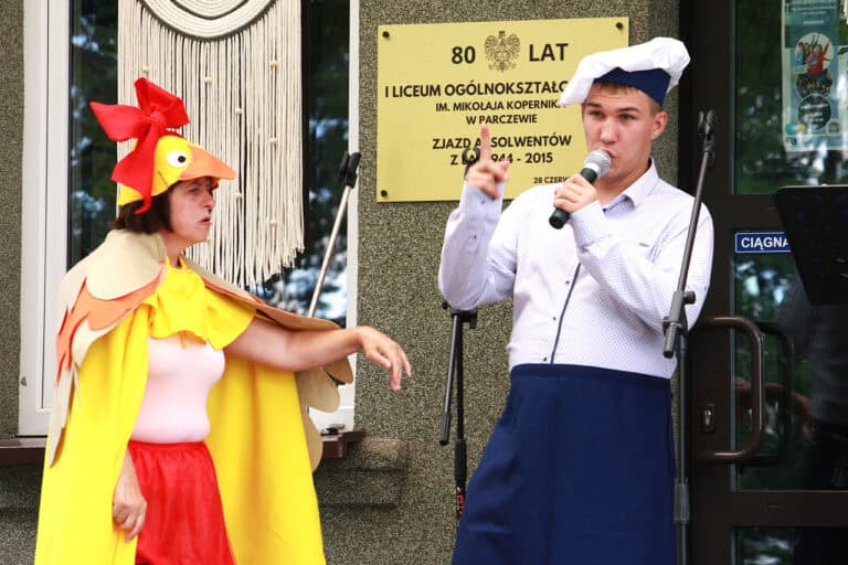 Od lewej kobieta w przebraniu kaczki, z dużą czerwoną kokardą na głowie wykonuje gest dłonią w stronę mężczyzny przebranego za kucharza. Świadczy o tym granatowy fartuch i biało granatowa czapka kucharza. Mężczyzna mówi coś do mikrofonu, patrząc przed siebie, unosi w górę prawą rękę i wskazuje palcem w górę