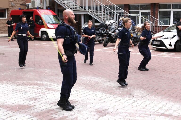 W kadrze grupa policjantów podczas skakania na skakankach. W tle widać motocykle, a w lewym górnym rogu bus Straży Pożarnej