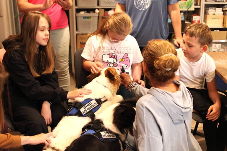 Cztery osoby pochylone nad psimi terapeutami. Jeden z nich opiera łapy o kolana dziewczynki. Dzieci głaszczą go