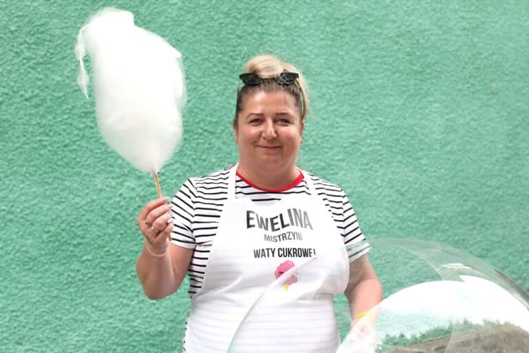 W centralnej części kadru uśmiechnięta kobieta (sołtys Sołectwa Milanów) prezentuje watę cukrową. Ma na sobie koszulkę w paski i biały fartuch z napisem: „Ewelina Mistrzyni Waty Cukrowej”