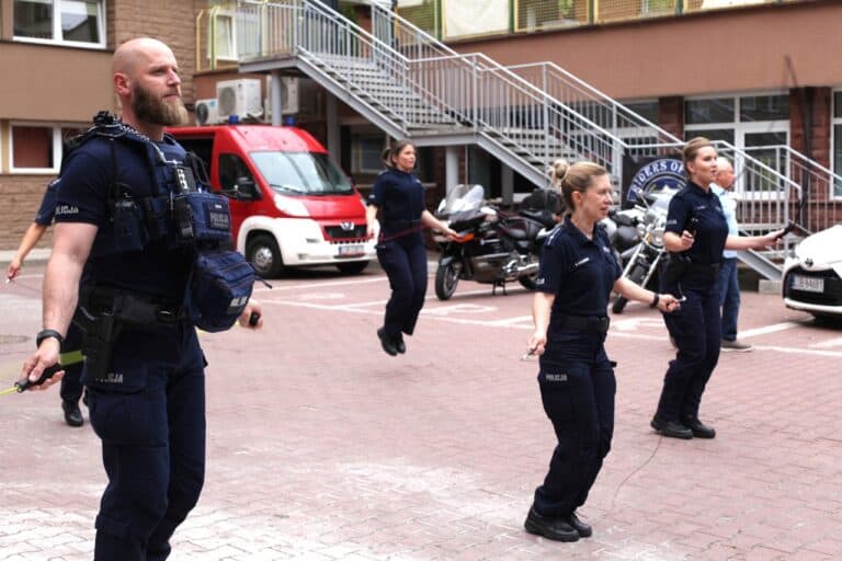 Grupa policyjna (trzy kobiety i mężczyzna) podczas skakania na skakankach. W tle stoją zaparkowane motocykle a w lewym górnym rogu bus Straży Pożarnej