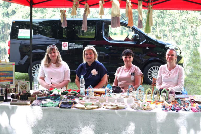 Stoisko Caritas Diecezji Siedleckiej. Do fotografii pozują cztery patrzące wprost do obiektywu kobiety. Siedzą za stołem z wyrobami rękodzielniczymi. W tle bus Warsztatu Terapii Zajęciowej w Parczewie