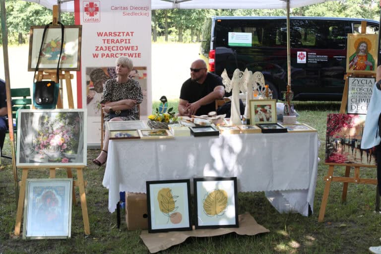 Stół z wyrobami rękodzielniczymi. Siedzą przy nim: kobieta i mężczyzna. Po lewej baner Caritas Diecezji Siedleckiej, WTZ w Parczewie