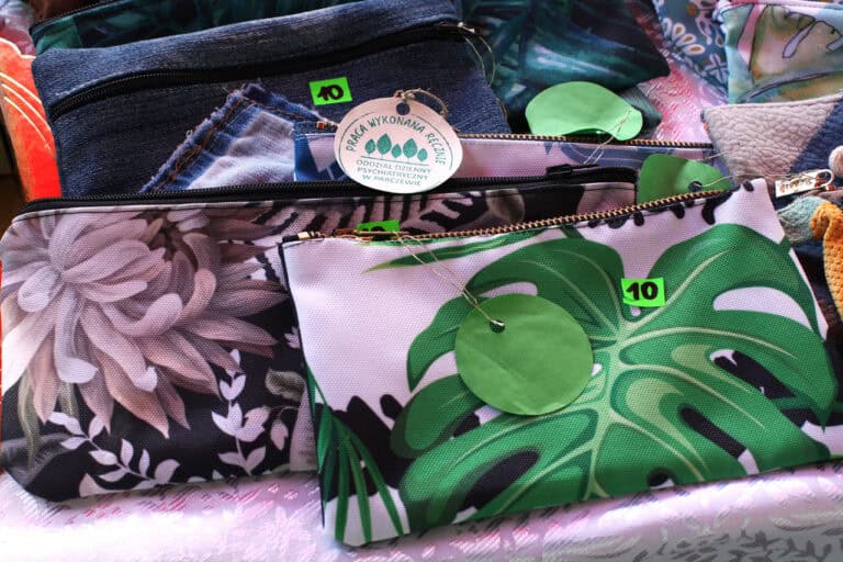 Zbliżenie na wyroby rękodzielnicze (etui, kosmetyczki w kwieciste i liściaste wzory). Do jednej z nich jest przyczepiona okrągła metka z napisem: Praca wykonana ręcznie. Oddział Dzienny Psychiatryczny w Parczewie