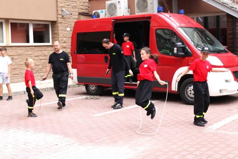 Na pierwszym planie dzieci i dorośli z ekipy strażaków, wszyscy w ruchu, ubrani w czerwone koszulki i czarne spodnie strażackie. Dziewczynka uchwycona w trakcie skoku. W tle mały czerwony wóz strażacki