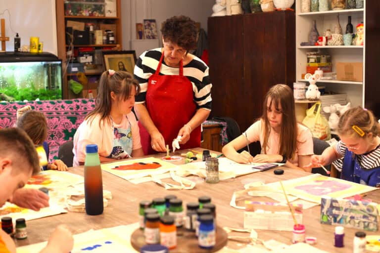 Fragment stołu, przy którym siedzą trzy dziewczynki. Nad nimi pani, która pomaga w pracach plastycznych. W tle wnętrze pracowni artystycznej, regały z rękodziełem