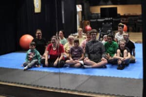 Grupa uczestników z instruktorką siedzą przed lustrem na niebieskiej macie. Część osób uśmiecha się. W lustrze odbija się też wnętrze sali i duże pomarańczowe piłki