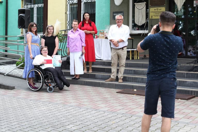 Fotografia grupowa z wręczenia nagrody uczestniczce wydarzenia. Z prawej strony kadru mężczyzna robiący zdjęcie