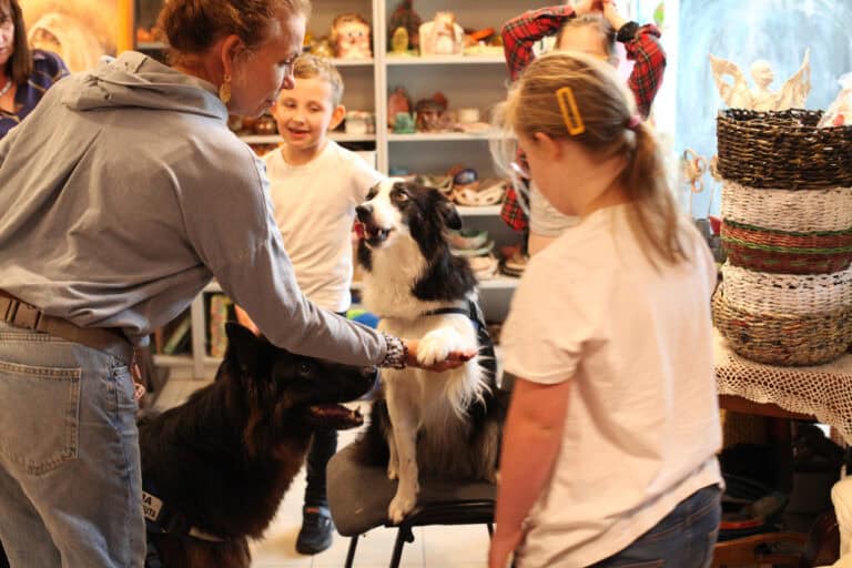 W centrum kadru na krześle siedzi Megi – pies terapeuta. Podaje łapę prowadzącej zajęcia. Wokół nich zgromadzone dzieci