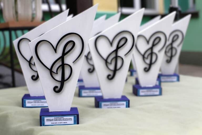 Zbliżenie na statuetki. Mają formę figury z graficznym motywem nuty wpisanej w serce. Czarny motyw na białym tle. Na niebieskiej podstawie plakietka z napisem: „IX Powiatowy Przegląd Twórczości Artystycznej Seniorów i Osób Niepełnosprawnych. Parczew, 13 lipca 2025 r.”