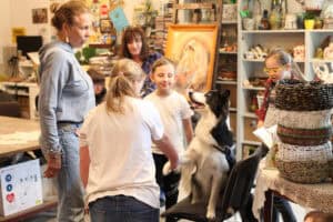 W centrum kadru na krześle Megi podająca łapę stojącej przed nią dziewczynce. Nad nimi prowadząca zajęcia Violetta Rycaj-Lewandowska i inne dzieci. W tle wnętrze pracowni