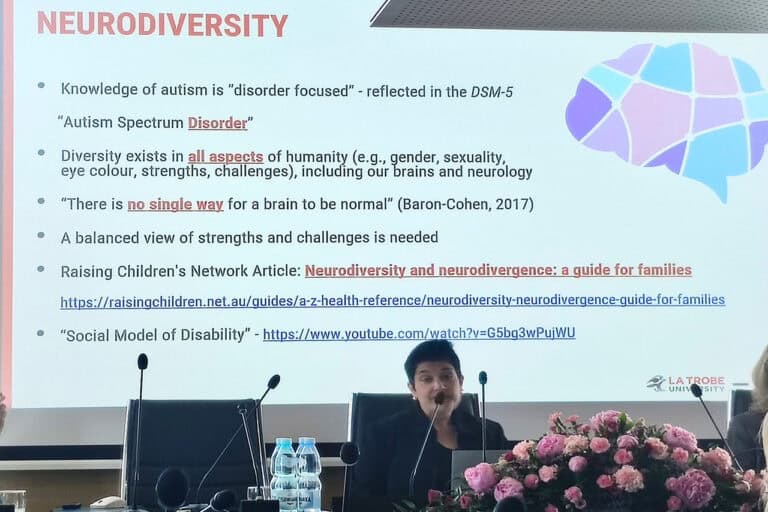 prof. Barbaro siedząca przy stole konferencyjnym, podczas prelekcji. Przed kobietą leży duży bukiet różowych kwiatów. Na ekranie wyświetlony slajd z treścią w języku angielskim pt. „Neuroróżnorodność” (tłumaczenie z ang.)