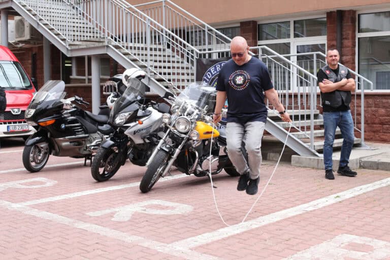 Na tle trzech motocykli mężczyzna uchwycony w ruchu (skacze na skakance). Obserwuje go drugi mężczyzna, znajdujący się po prawej stronie kadru