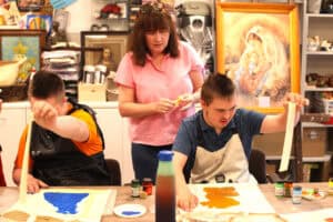 Przy stole dwóch chłopców. Obaj odrywają taśmę malarską ze swoich prac. Nad nimi stoi kobieta. W tle komody z obrazami