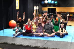 Wnętrze sali z lustrem, w którym widać grupę młodzieży. Niektórzy z nich radośnie unoszą ręce w górę. Siedzą na niebieskiej macie. Na ścianie za nimi widać półkę z piłkami. Obok grupy, na macie leży duża pomarańczowa piłka