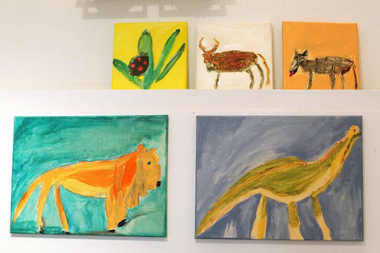 Widok na jedną ze ścian galerii z pięcioma obrazami różnej wielkości, wyeksponowanymi w dwóch rzędach. Obrazy przedstawiają zwierzęta namalowane w intensywnych barwach