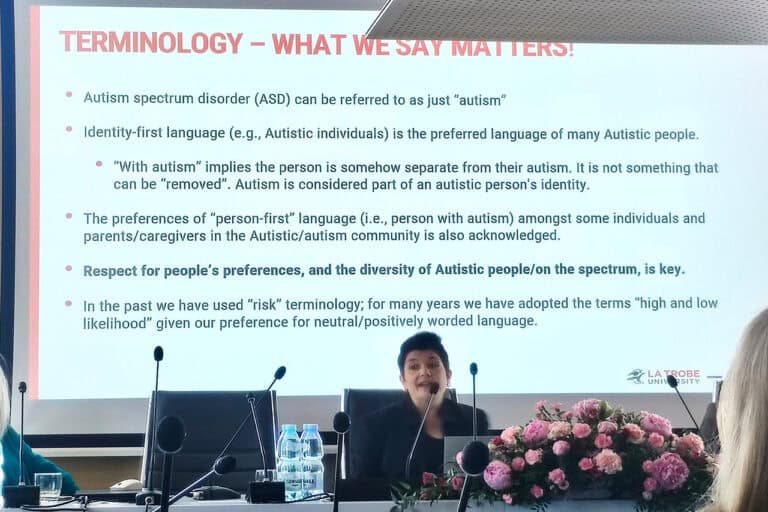 prof. Barbaro siedząca przy stole konferencyjnym, podczas prelekcji. Przed kobietą leży duży bukiet różowych kwiatów. Za nią ekran z wyświetlonym anglojęzycznym slajdem pt. „Terminologia – to co mówimy ma znaczenie” (tłum. z angielskiego)