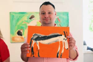 W centralnej części kadru Adam Saran pokazujący do obiektywu jeden ze swoich obrazów. W tle, za artystą znajduje się kolejny obraz zawieszony na ścianie