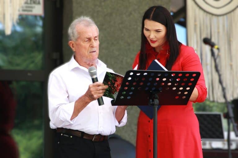 W kadrze od lewej: uczestnik przeglądu w kategorii seniorów, ubrany w białą koszulę, przegląda program wydarzenia. Towarzyszy mu młoda kobieta - prowadząca przegląd. Ma na sobie czerwoną sukienkę