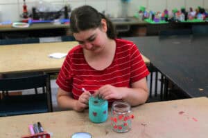 Dziewczynka ubrana w czerwoną bluzkę w białe poziome paski zdobi farbami słoik. Na jego błękitnym tle maluje czerwone serca. Za nią wnętrze pracowni artystycznej