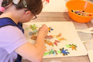 Dziewczynka uchwycona z profilu, pochylona nad płócienną torbą, którą zdobi farbami. Na torbie kolorowe motyle i kwiaty. W prawym górnym rogu pomarańczowa miska ze stemplami do farb