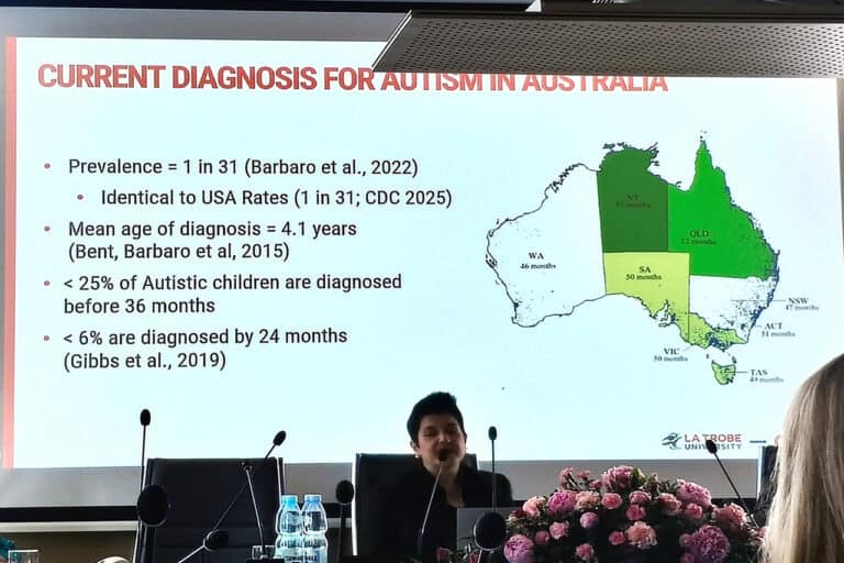prof. Barbaro siedząca przy stole konferencyjnym, podczas prelekcji. Przed kobietą leży duży bukiet różowych kwiatów. Tuż za nią monitor z mapą i danymi dotyczącymi aktualnej diagnozy autyzmu w Australii; treści zaprezentowano w języku angielskim