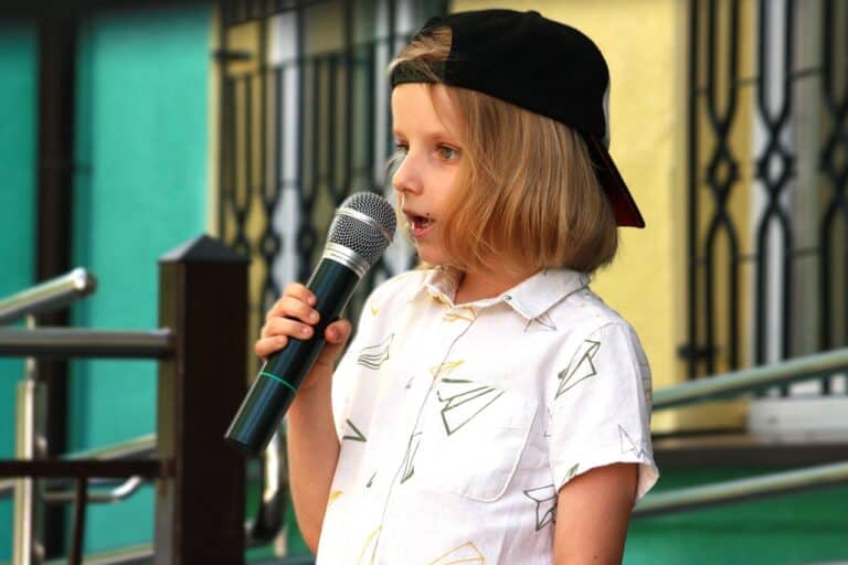Fotografia portretowa najmłodszego uczestnika przeglądu artystycznego. Chłopiec śpiewa do mikrofonu. Ma na sobie jasną koszulę w duże wzory i czapkę typu „kaszkietówka”, założoną daszkiem do tyłu