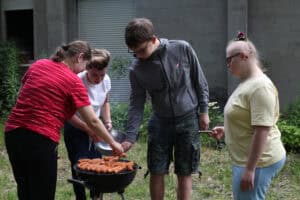 Na pierwszym planie grill z kiełbaskami. Wokół grilla cztery osoby. Trzy z nich przekładają kiełbaski z miski, jedna trzyma widelec i przygląda się tej scenie. W tle ściana budynku