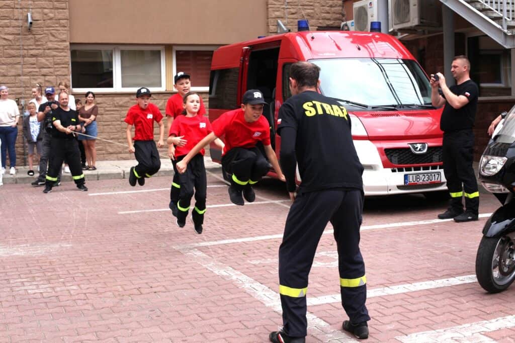 Ekipa dzieci i strażaków w skokach synchronicznych. Dwóch mężczyzn trzyma długą skakankę. Przeskakuje przez nią czworo dzieci. Wszystkie dzieci uchwycone podczas podskoku