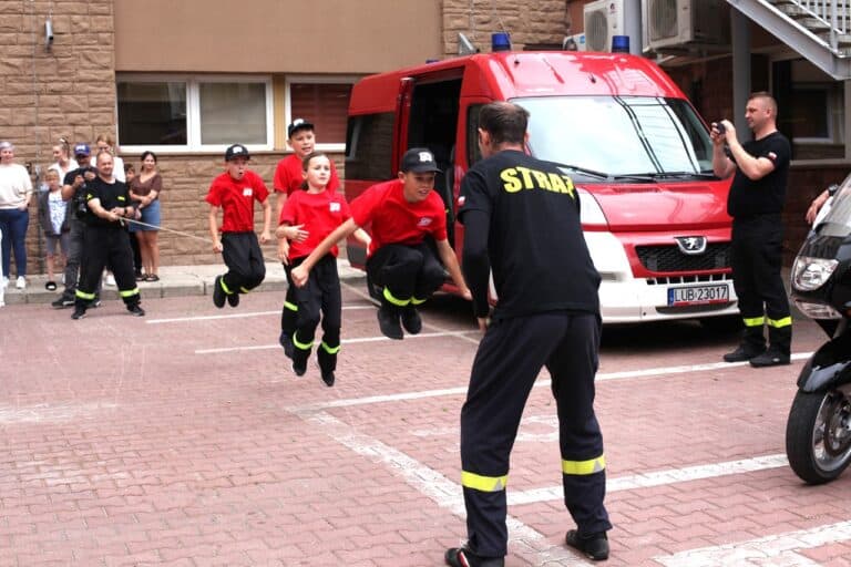 Ekipa dzieci i strażaków w skokach synchronicznych. Dwóch mężczyzn trzyma długą skakankę. Przeskakuje przez nią czworo dzieci. Wszystkie dzieci uchwycone podczas podskoku