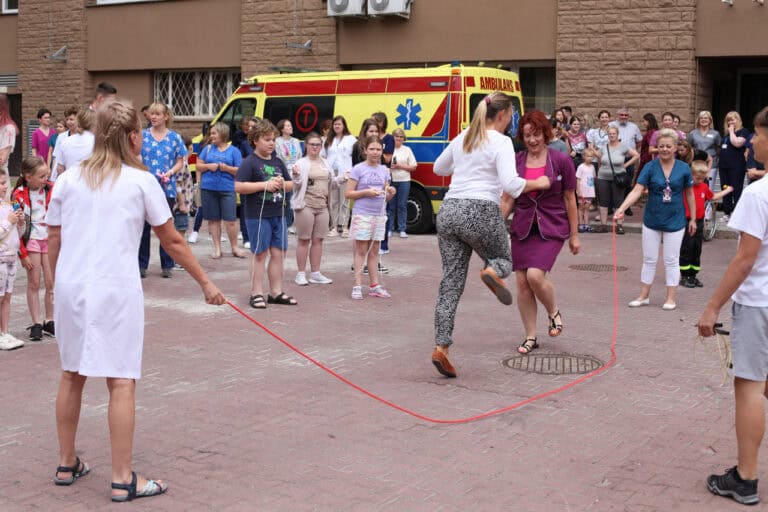 Kadr wypełniony uczestnikami akcji. Na pierwszym planie dwie kobiety z personelu medycznego skaczą przez długą skakankę trzymaną przez ich koleżanki. Dookoła nich obserwatorzy: dzieci, młodzież i dorośli