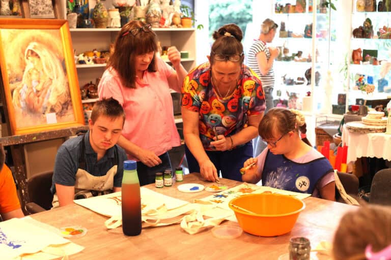 Przy stole siedzą chłopiec i dziewczynka nad swoimi pracami artystycznymi. Pomagają im stojące obok dwie kobiety (wśród nich pani Małgosia). Po lewej stronie kadru obraz na sztalugach, w tle wnętrze pracowni