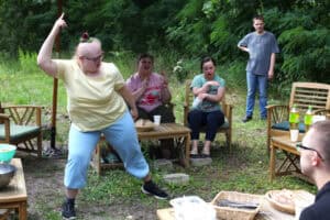 Na tle zieleni krzesła i stolik. Na pierwszym planie z lewej młoda kobieta tańcząc unosi w górę prawą rękę. Tuż za nią, w centrum kadru, dwie młode kobiety siedząc na krzesłach poruszają rękami w rytm muzyki. Za nimi, stoi młody mężczyzna z ręką podpartą na biodrze