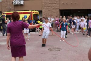 Na pierwszym planie chłopiec skacze na skakance trzymanej przez dwie kobiety z personelu medycznego. W tle uczestnicy akcji. W lewym górnym rogu widać żółto czerwony ambulans