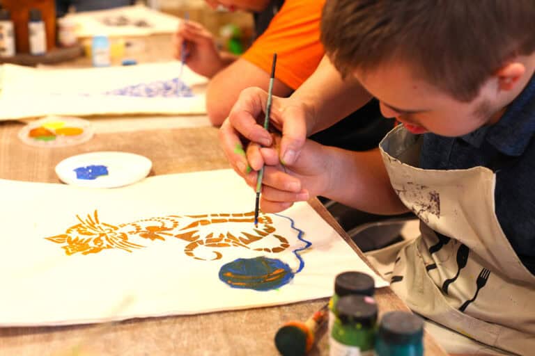 Chłopiec pochylony nad swoją pracą plastyczną. Zbliżenie na dłonie chłopca i opiekunki, która wspiera go w malowaniu. Ich dłonie trzymają pędzel nad torbą z ilustracją kota i kłębka włóczki