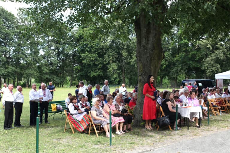Publiczność w plenerze w otoczeniu drzew. Od lewej grupa seniorów. Publiczność siedzi na ławkach, część stoi w grupach. Po prawej stronie stolik komisji sędziowskiej