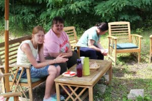 Trzy młode kobiety siedzą na ławce, przy stoliku z jedzeniem. Dwie z nich uśmiechają się patrząc w obiektyw, trzecia nachylona, patrzy w dół. W tle zieleń