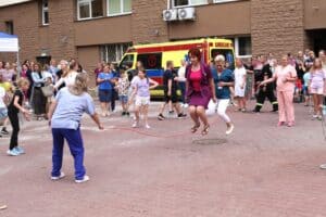 Na pierwszym planie cztery kobiety z personelu medycznego szpitala podczas zabawy ze skakanką. W tle uczestnicy akcji i ambulans w żółto czerwonych barwach